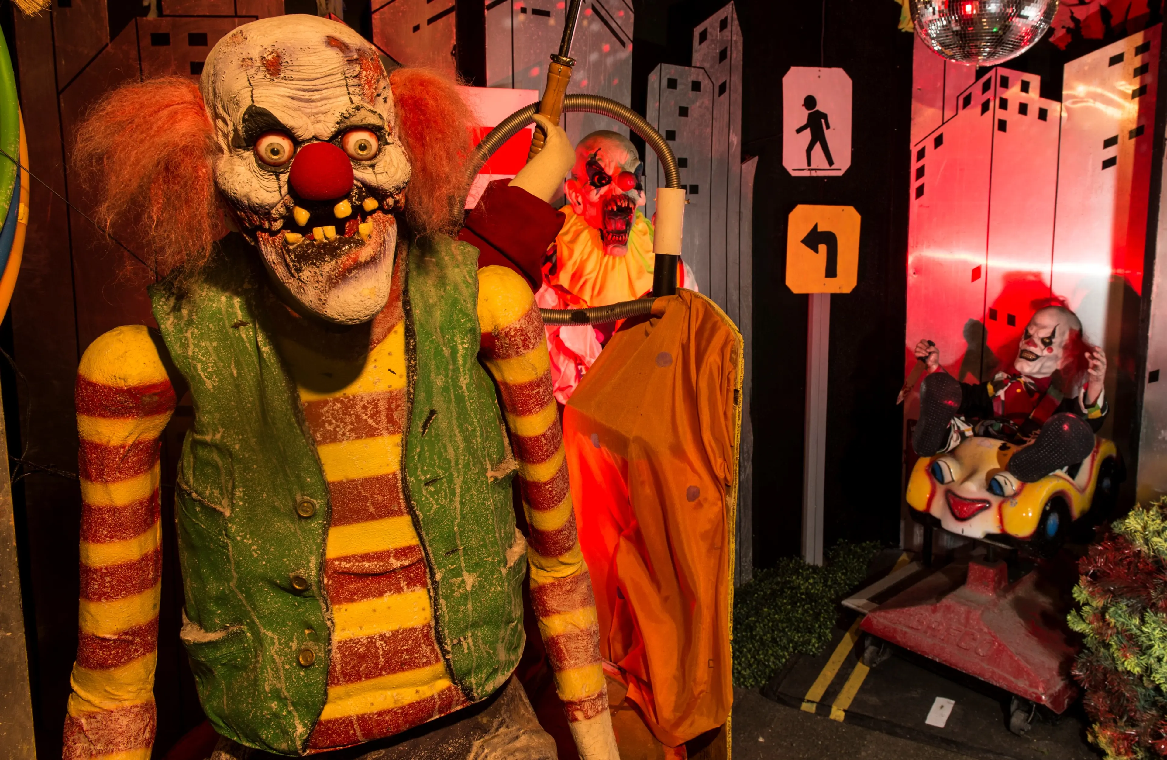 Three creepy clown figures in a dimly lit haunted house setting with cityscape backdrop and traffic signs.