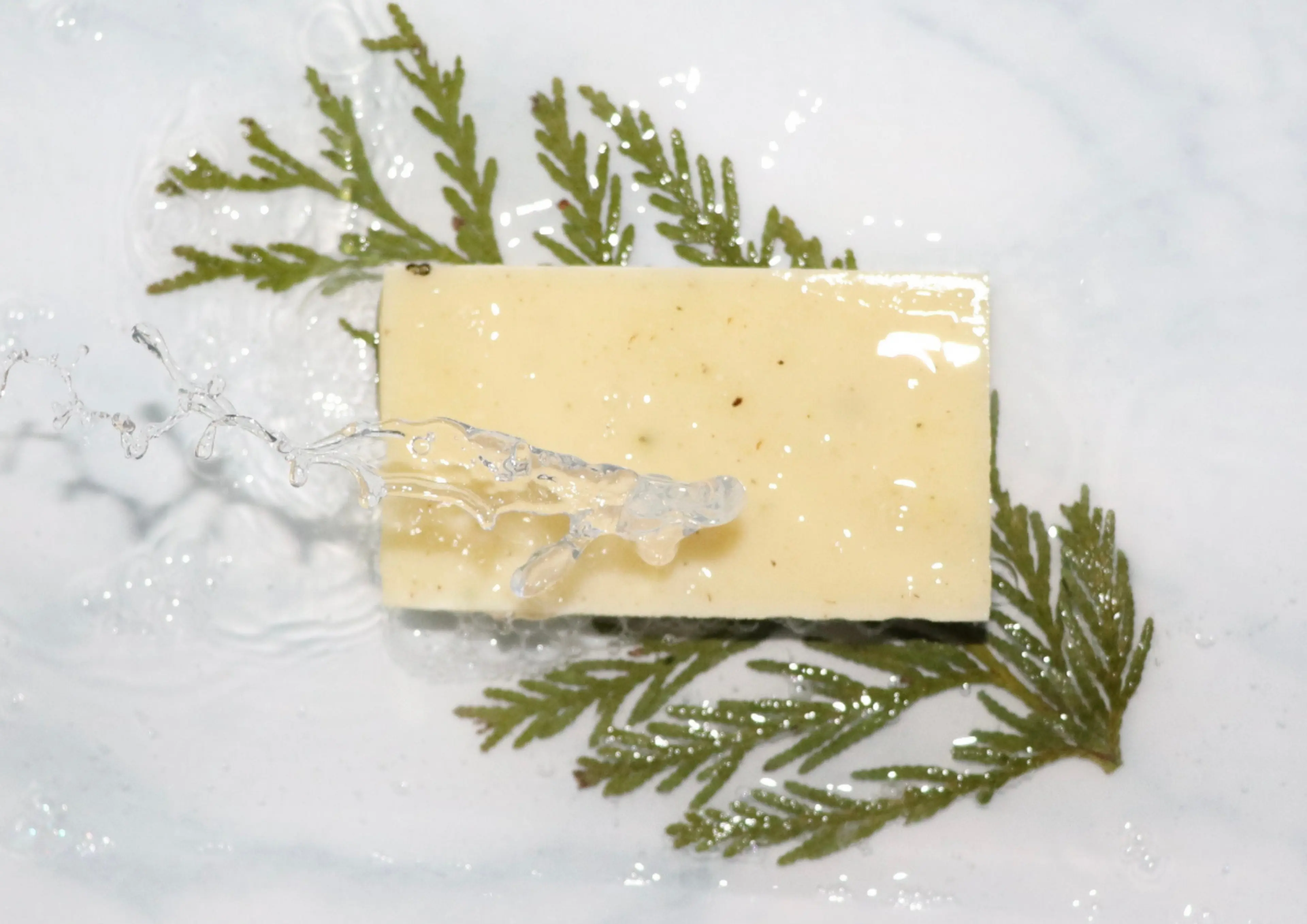 Yellow soap bar on green leaves with water splashing over it on a white surface.