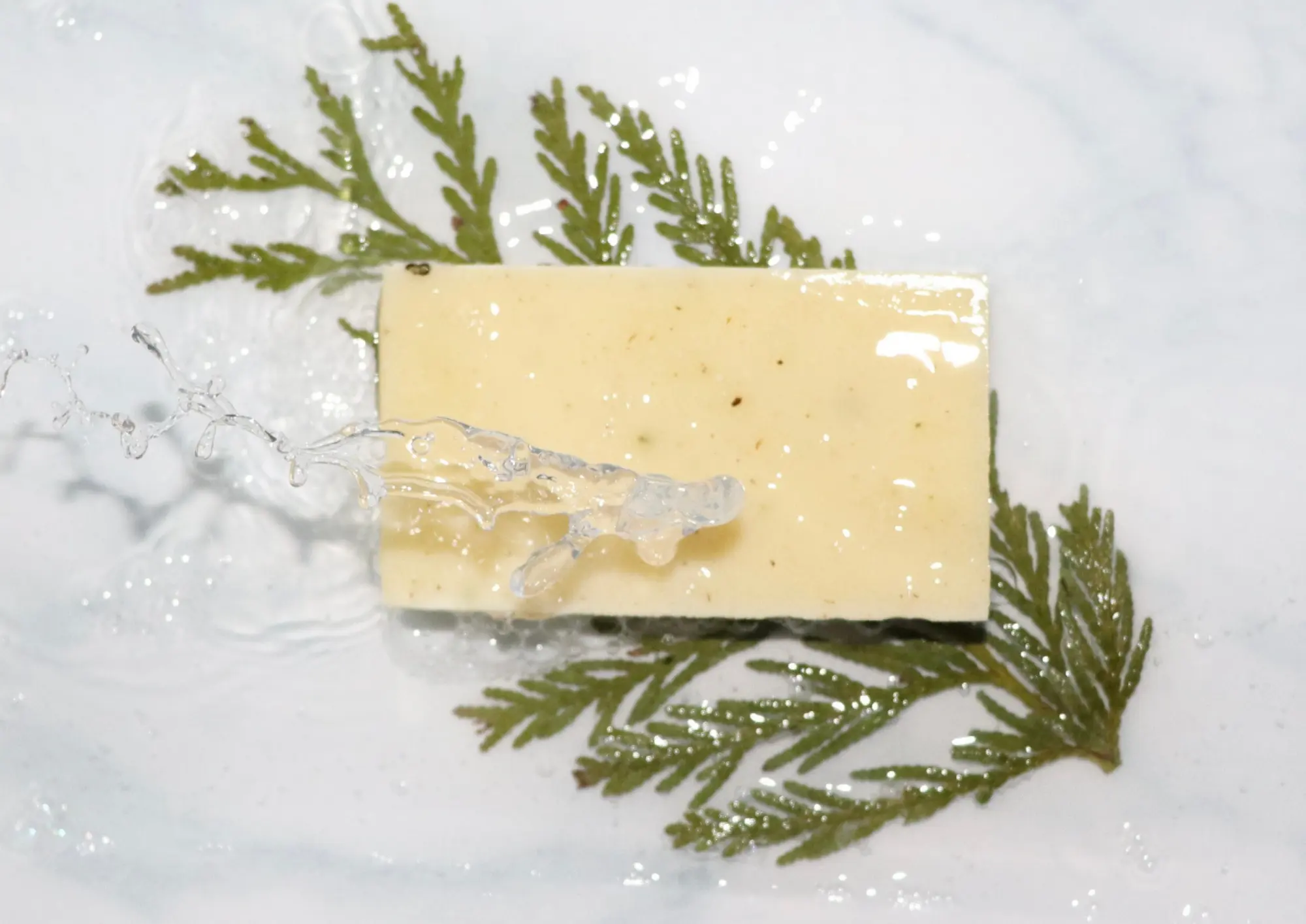 Yellow soap bar on green leaves with water splashing over it on a white surface.