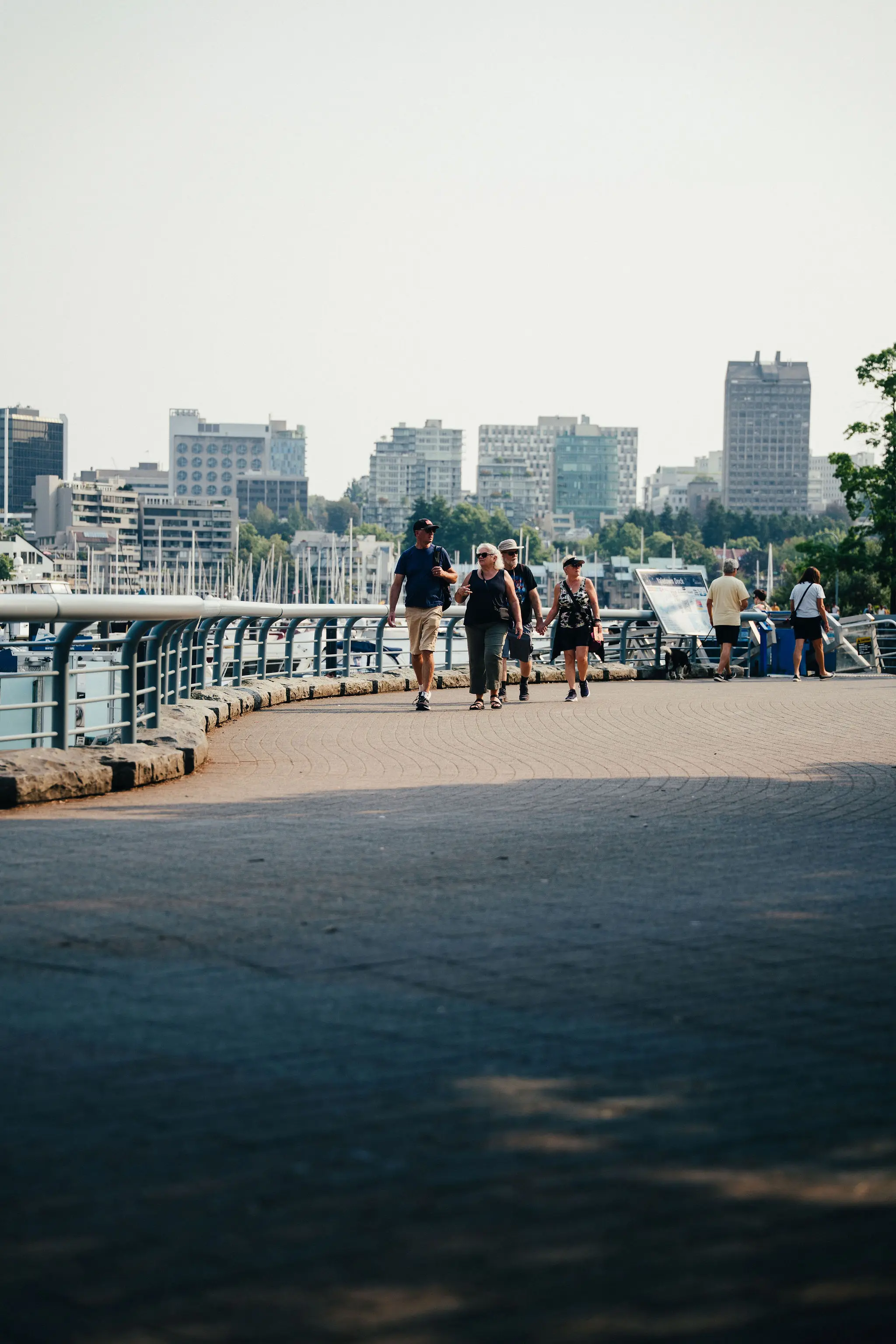 People walking on a curved waterfront promenade with city buildings and boats in the background.