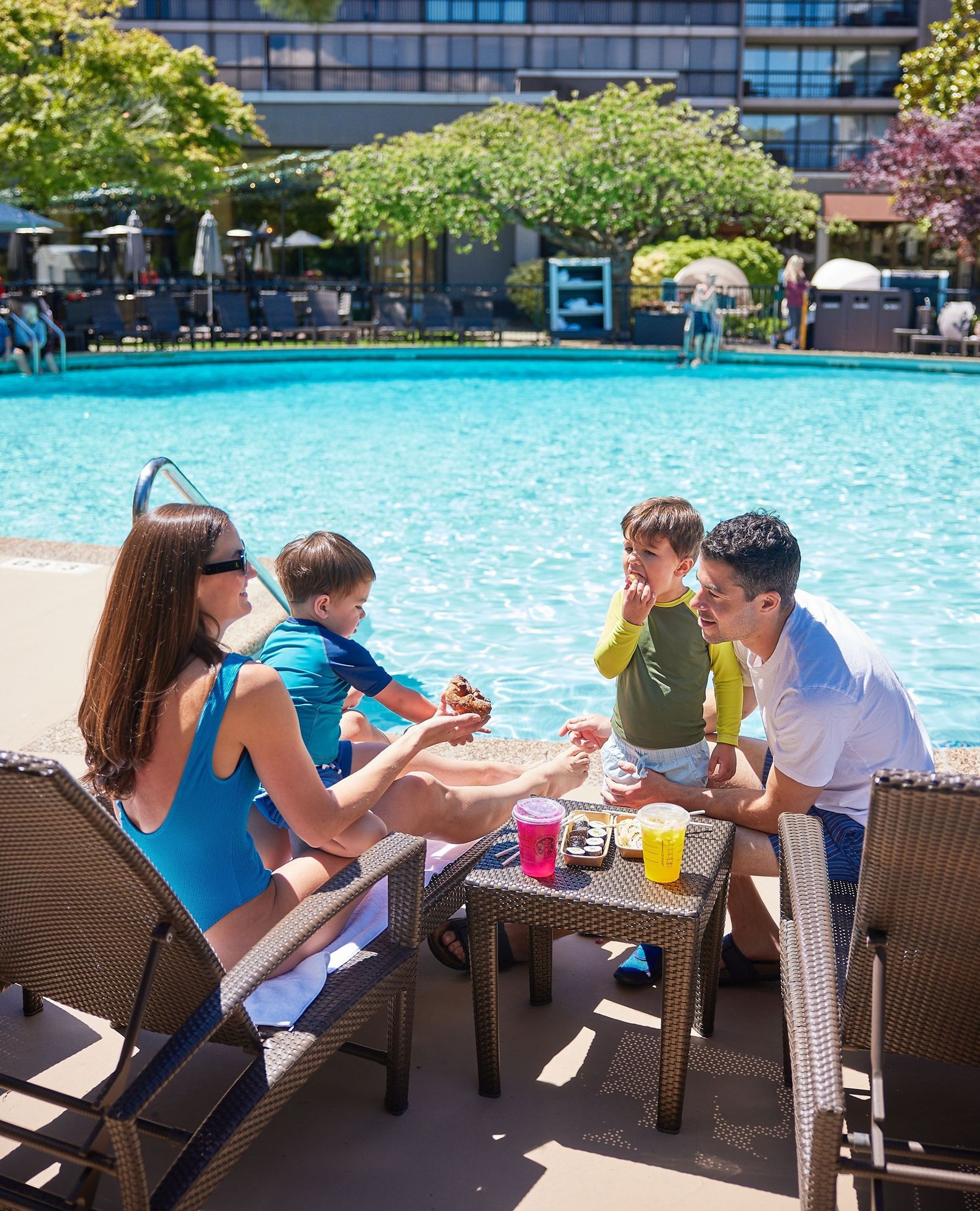 Family of four by a poolside with two children eating snacks and drinks on a small table.
