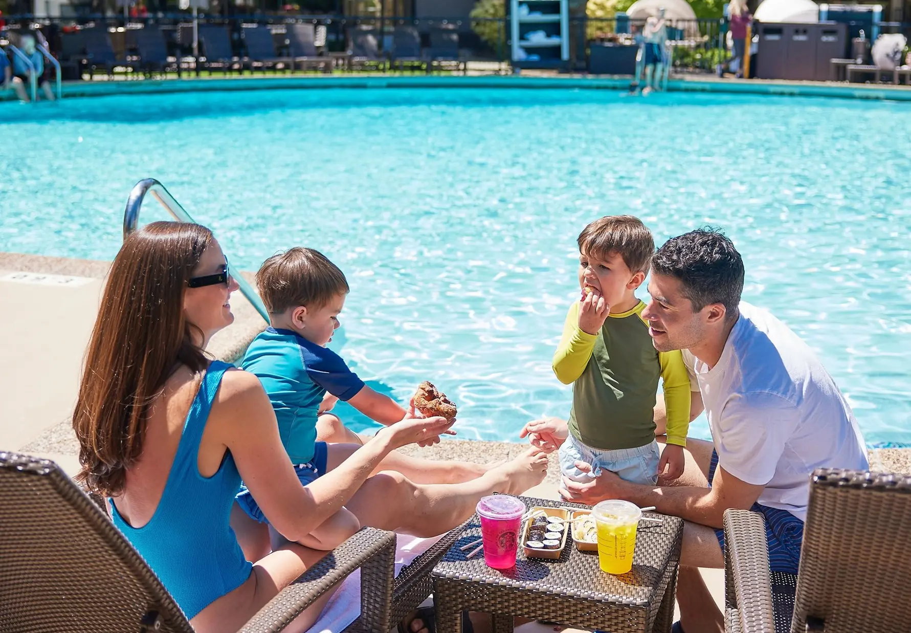 Family of four by a poolside with two children eating snacks and drinks on a small table.