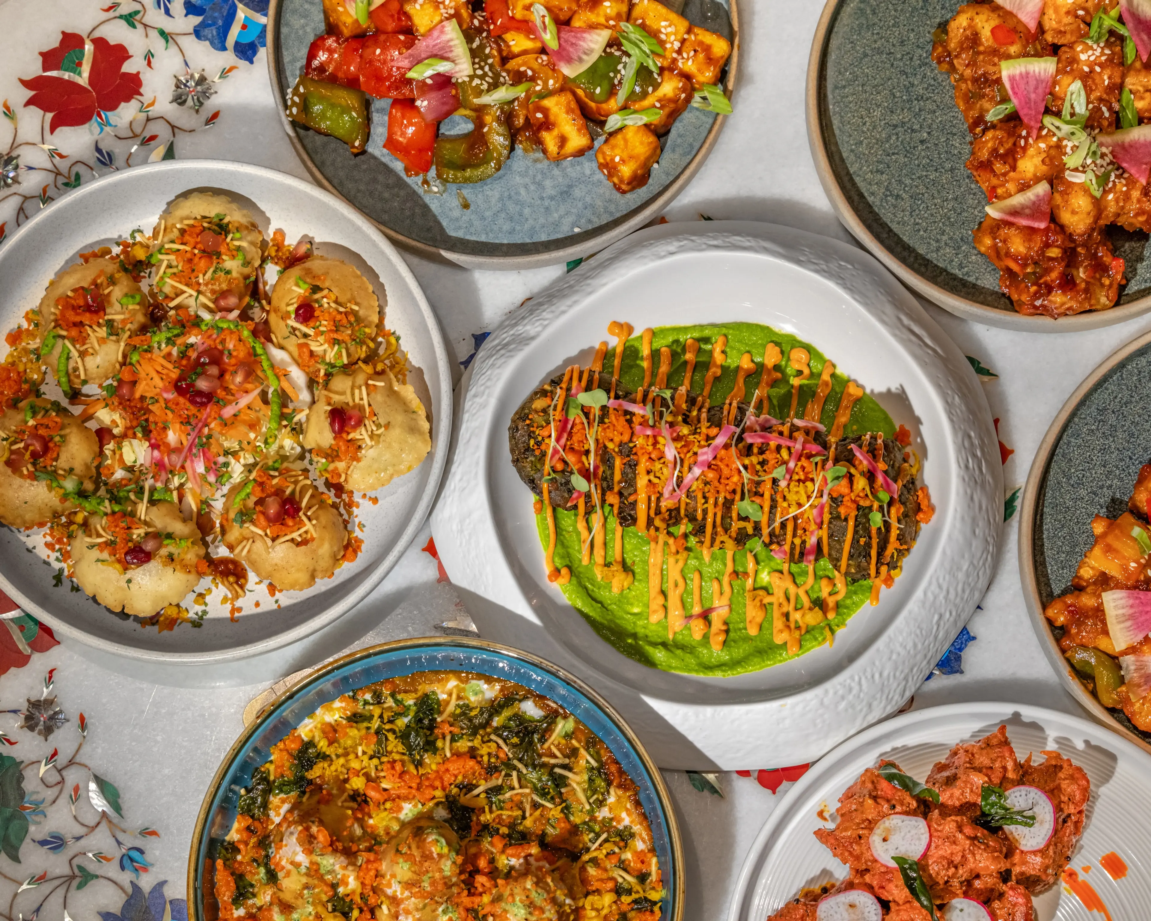 Top-down view of a vibrant spread of Indian dishes at Desi Indian Lounge, including gol gappa (pani puri), paneer tikka, and various colorful curries garnished with fresh herbs, radish, and pomegranate seeds. The plates are arranged on a decorative white tablecloth with a traditional floral pattern.