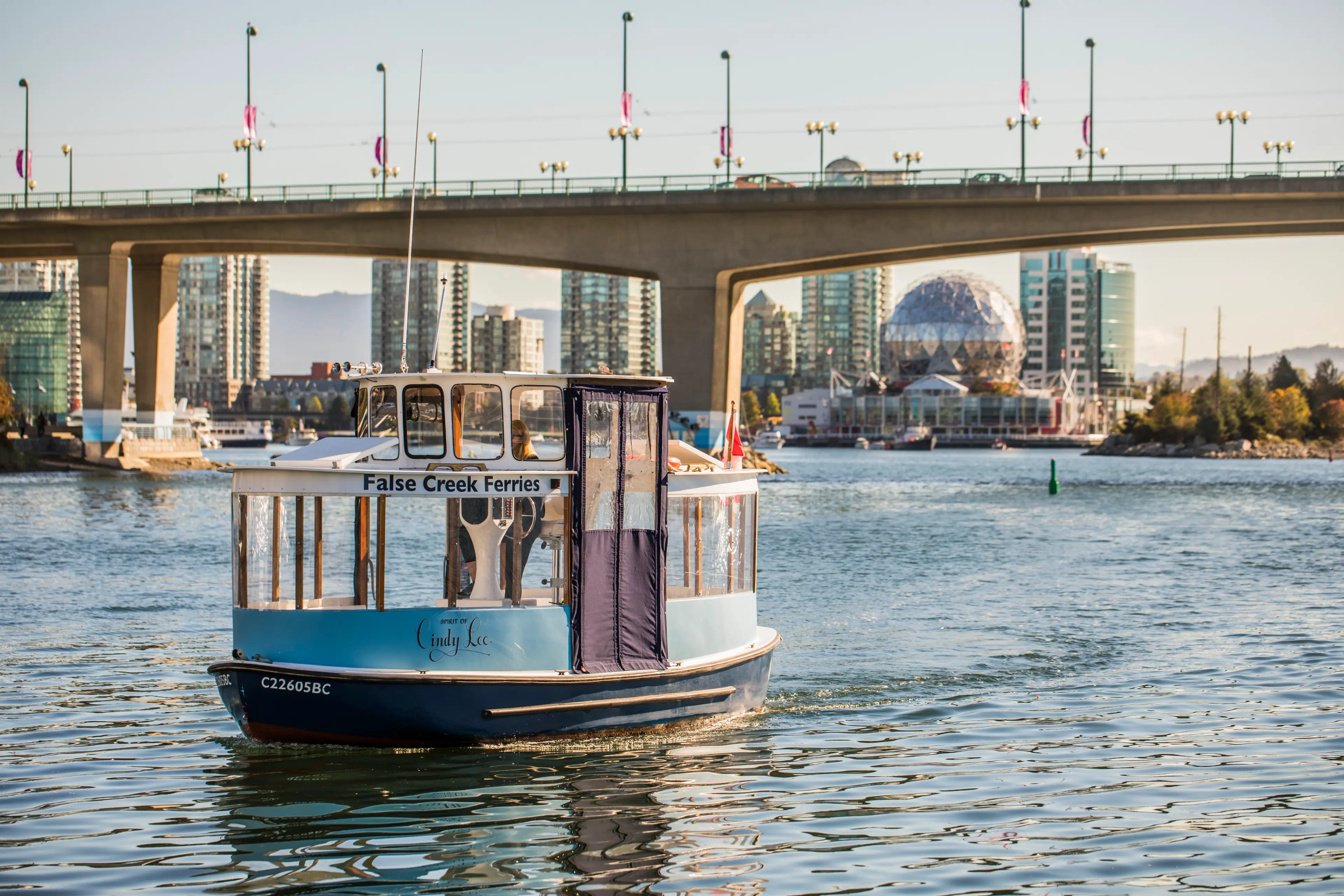 Small False Creek ferry boat on water with city buildings and a bridge in the background.