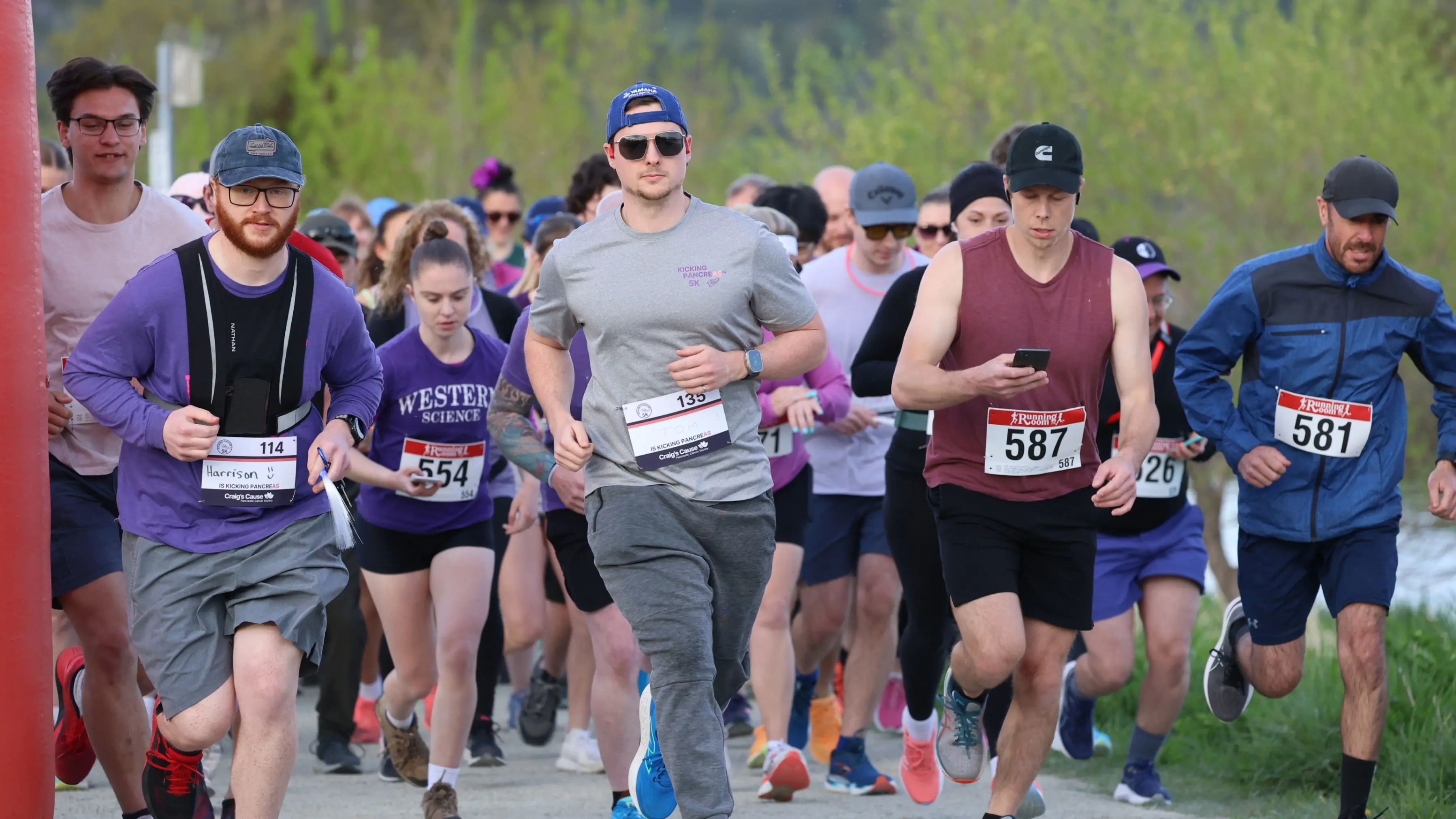 A group of runners wearing race bibs running outdoors on a path with greenery in the background.