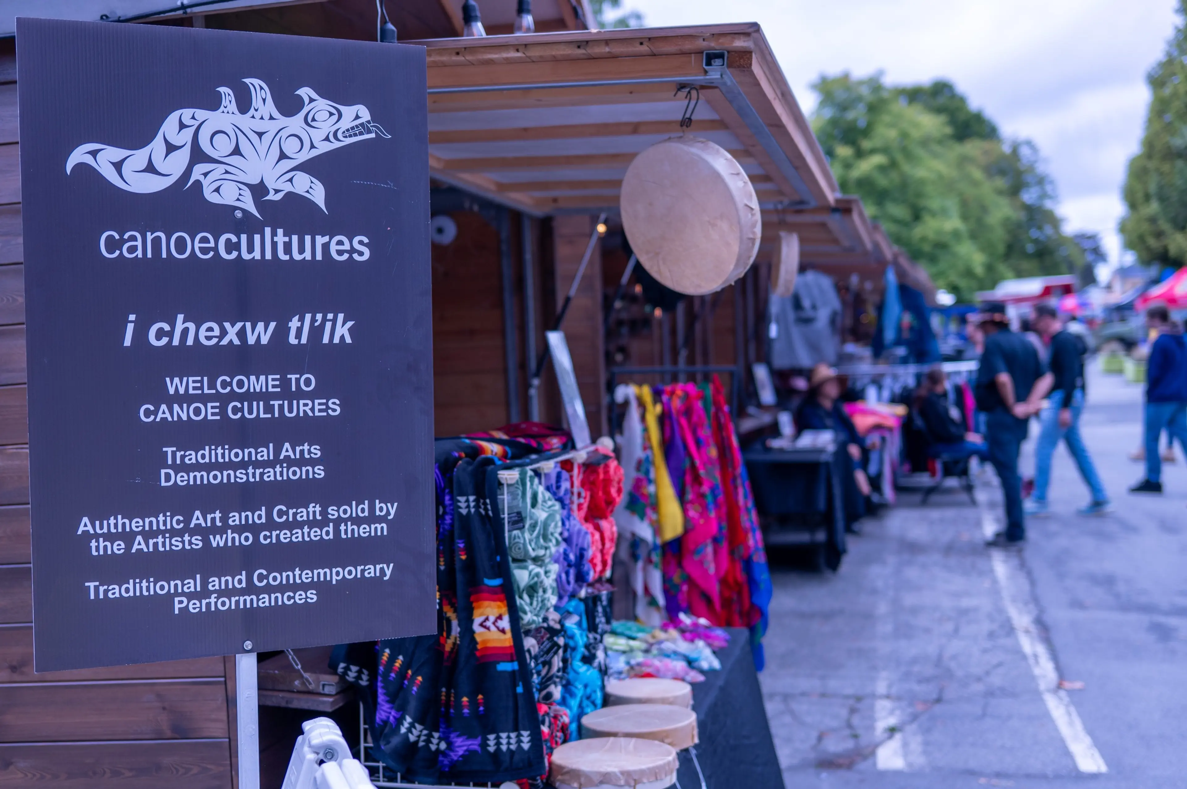 Sign for Canoe Cultures with traditional arts and crafts displayed at outdoor market stalls and people walking nearby.