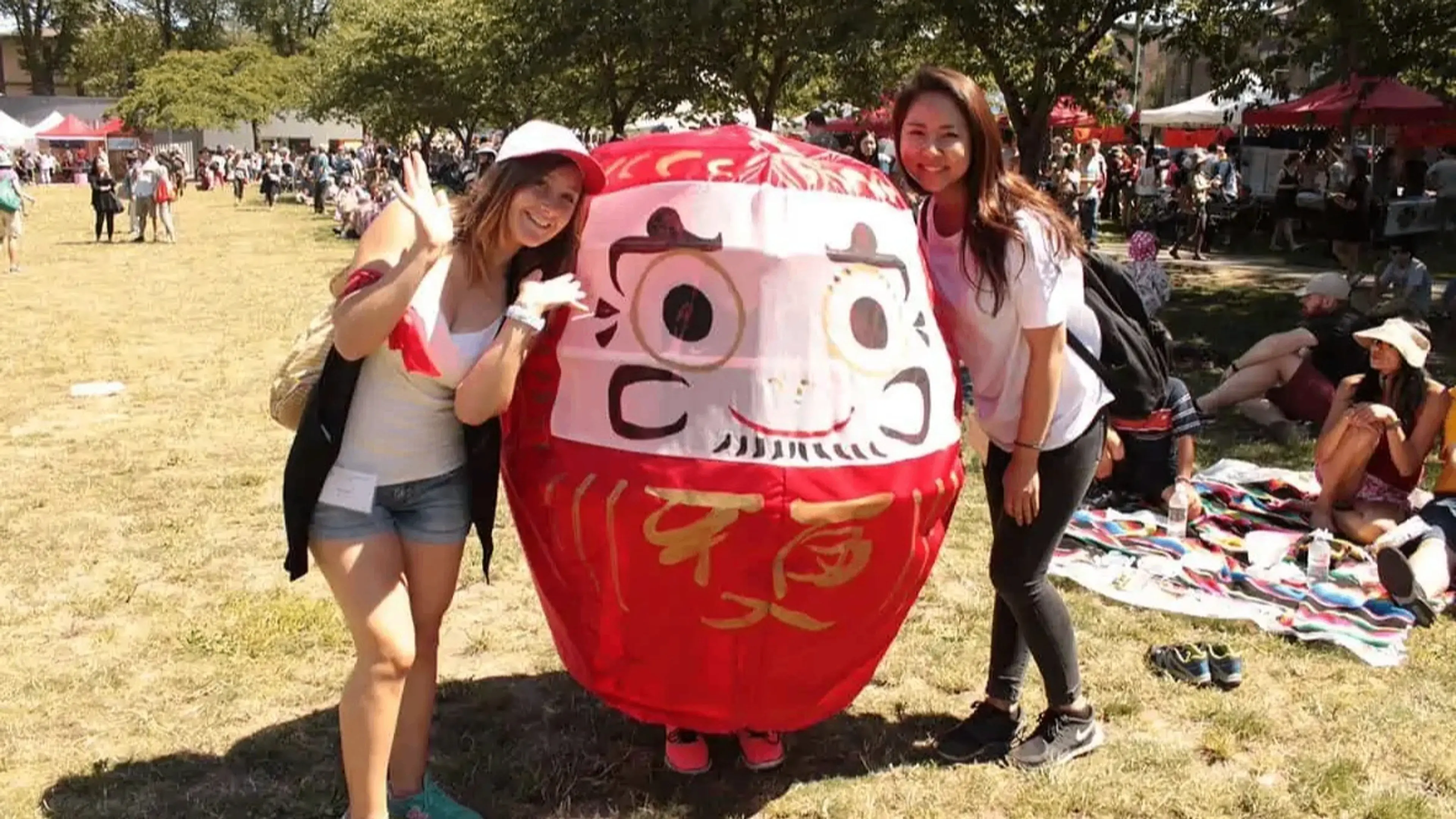 Two people posing and smiling beside a large red Daruma doll figure at an outdoor event with others sitting on blankets in the background.