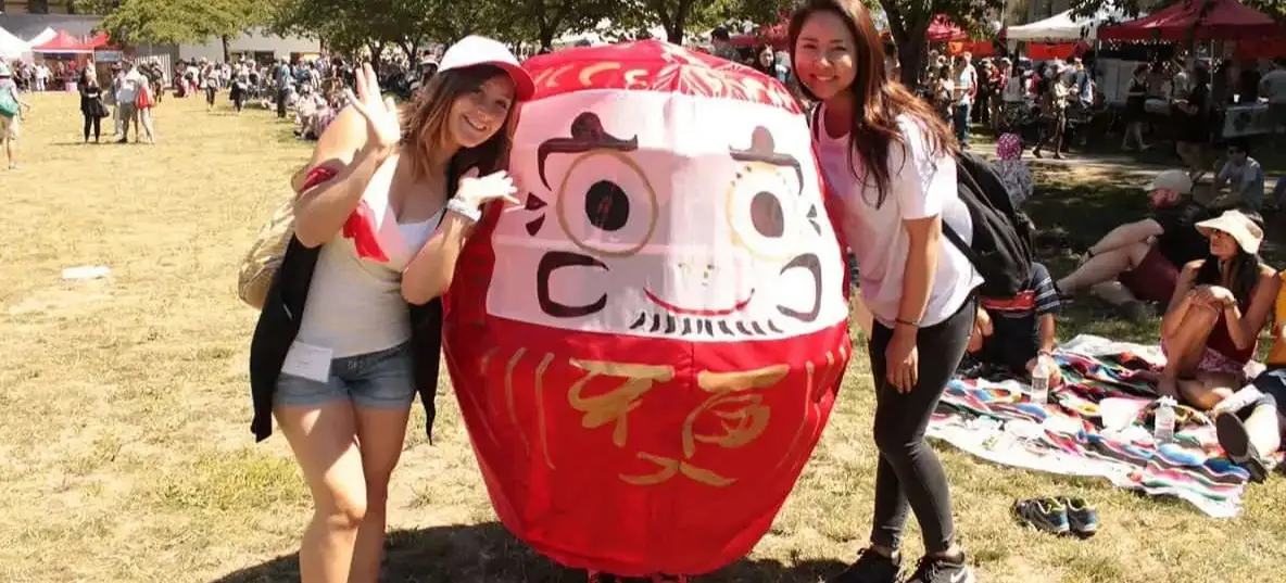 Two people posing and smiling beside a large red Daruma doll figure at an outdoor event with others sitting on blankets in the background.