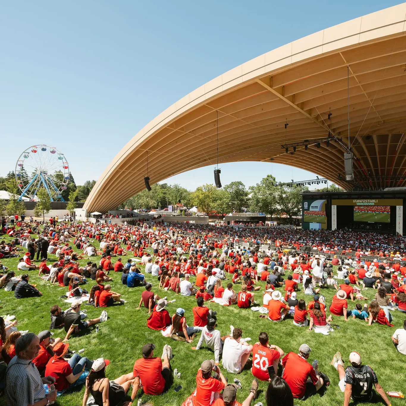 Large outdoor amphitheater with a wooden arched roof, a crowd sitting on grass, and a band playing on stage with screens showing musicians.