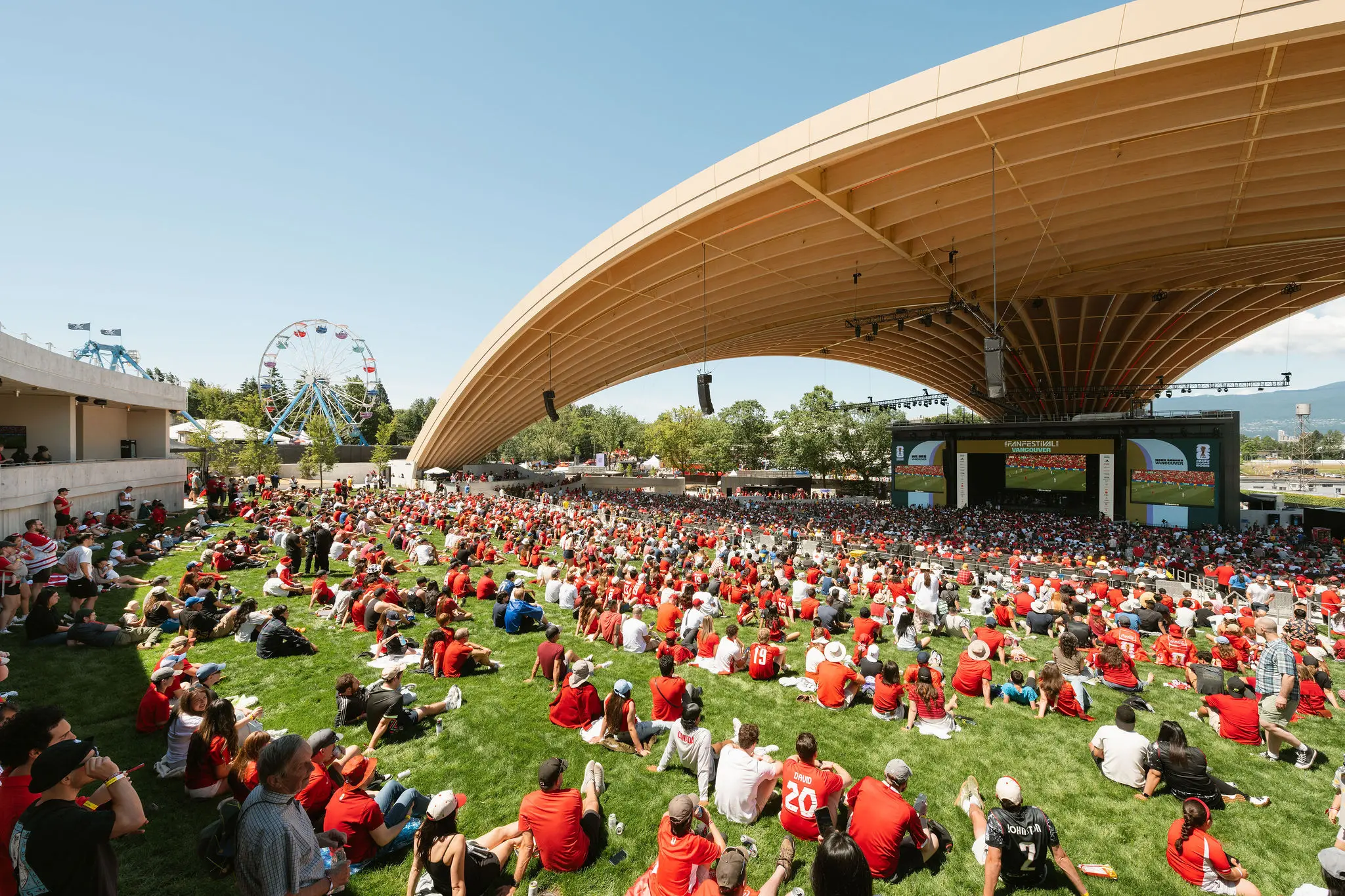 Large crowd wearing red sitting on grass under a curved wooden canopy watching a large screen with a Ferris wheel in the background.