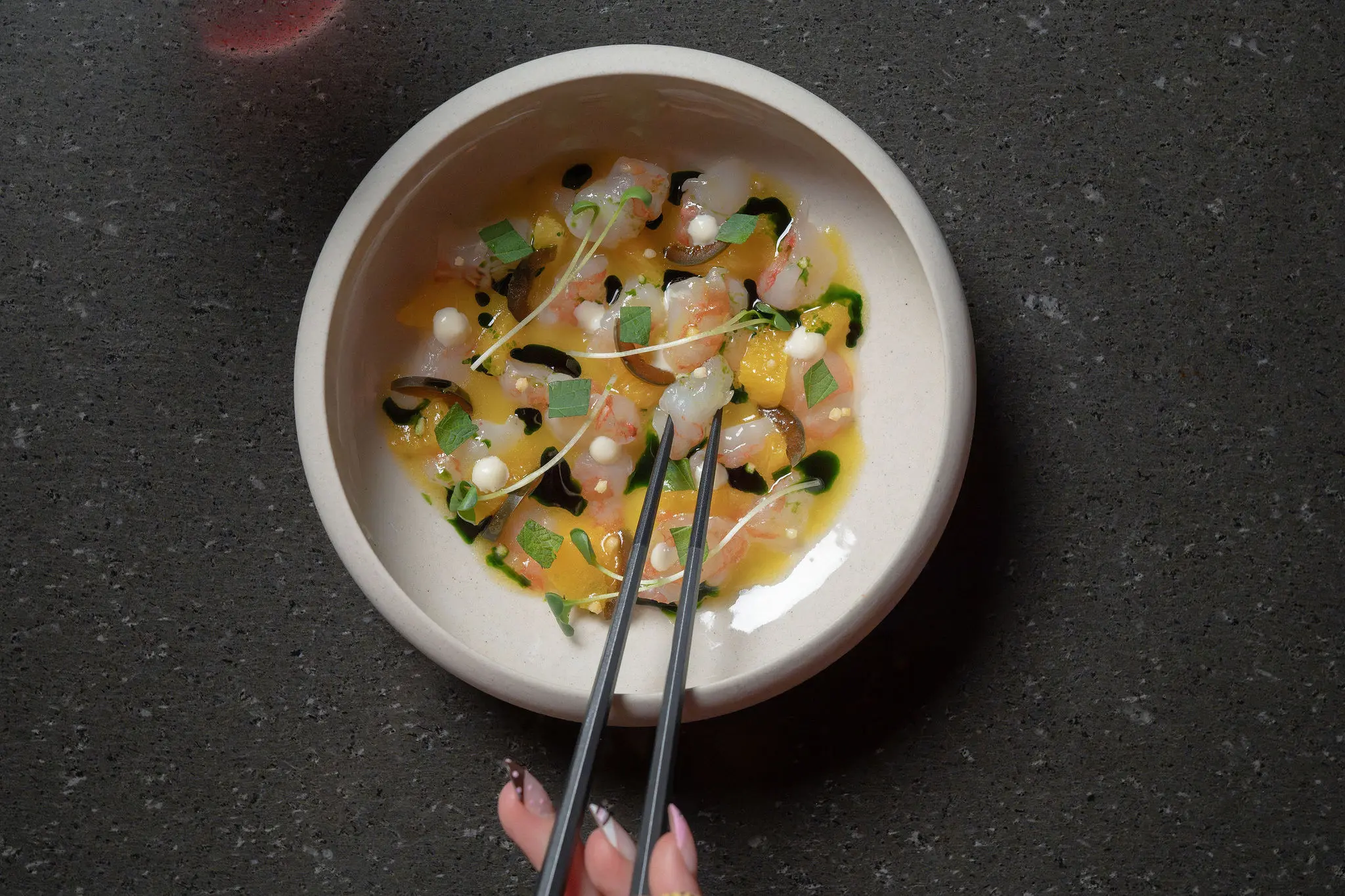 Hand holding chopsticks picking up food from a bowl with colorful garnished dish on a dark surface.