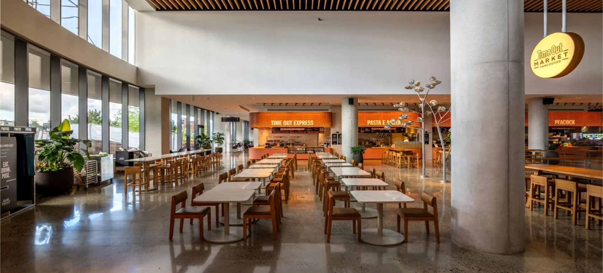 Empty modern food court with rows of tables and chairs, large windows, and illuminated food stall signs.