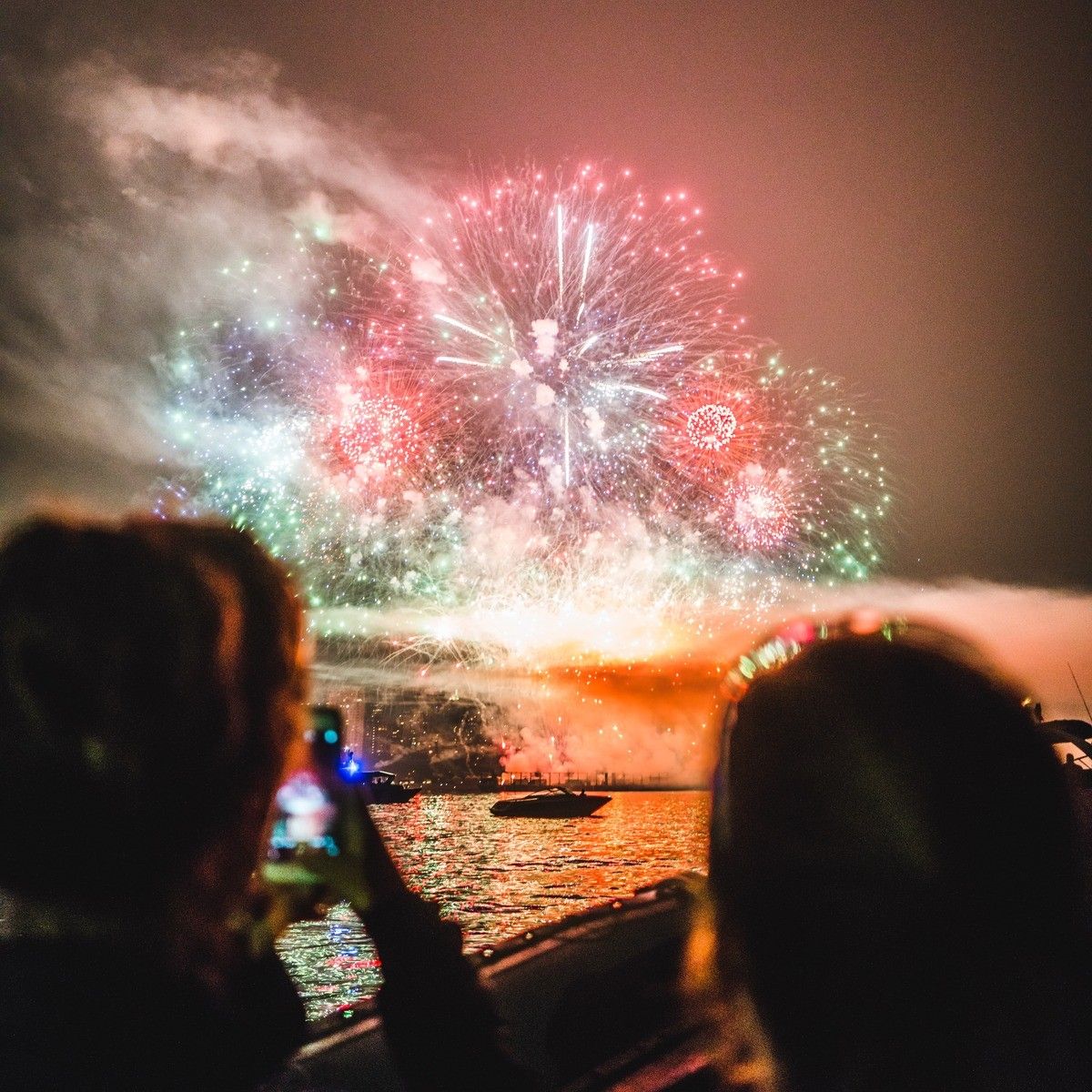 Two people on a boat watching and recording colorful fireworks over water at night.