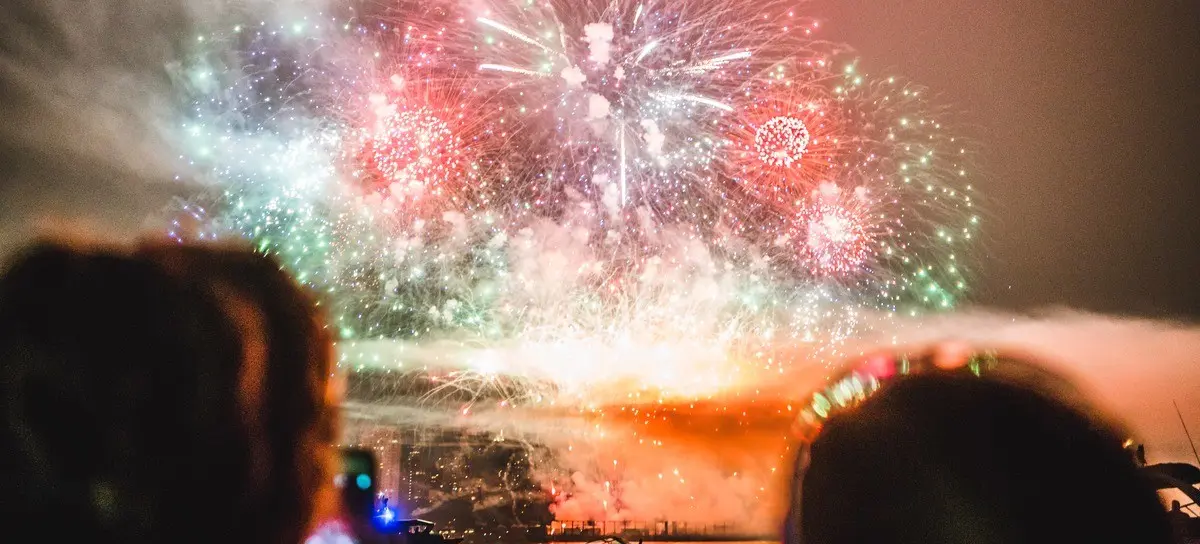 Two people on a boat watching and recording colorful fireworks over water at night.