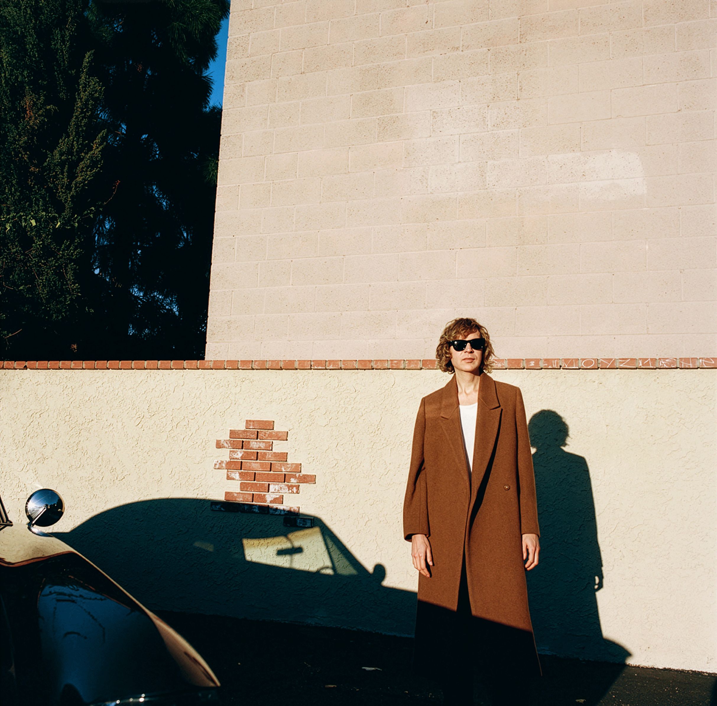 Person wearing brown coat and sunglasses standing against a beige wall with a shadow of a car in the foreground.