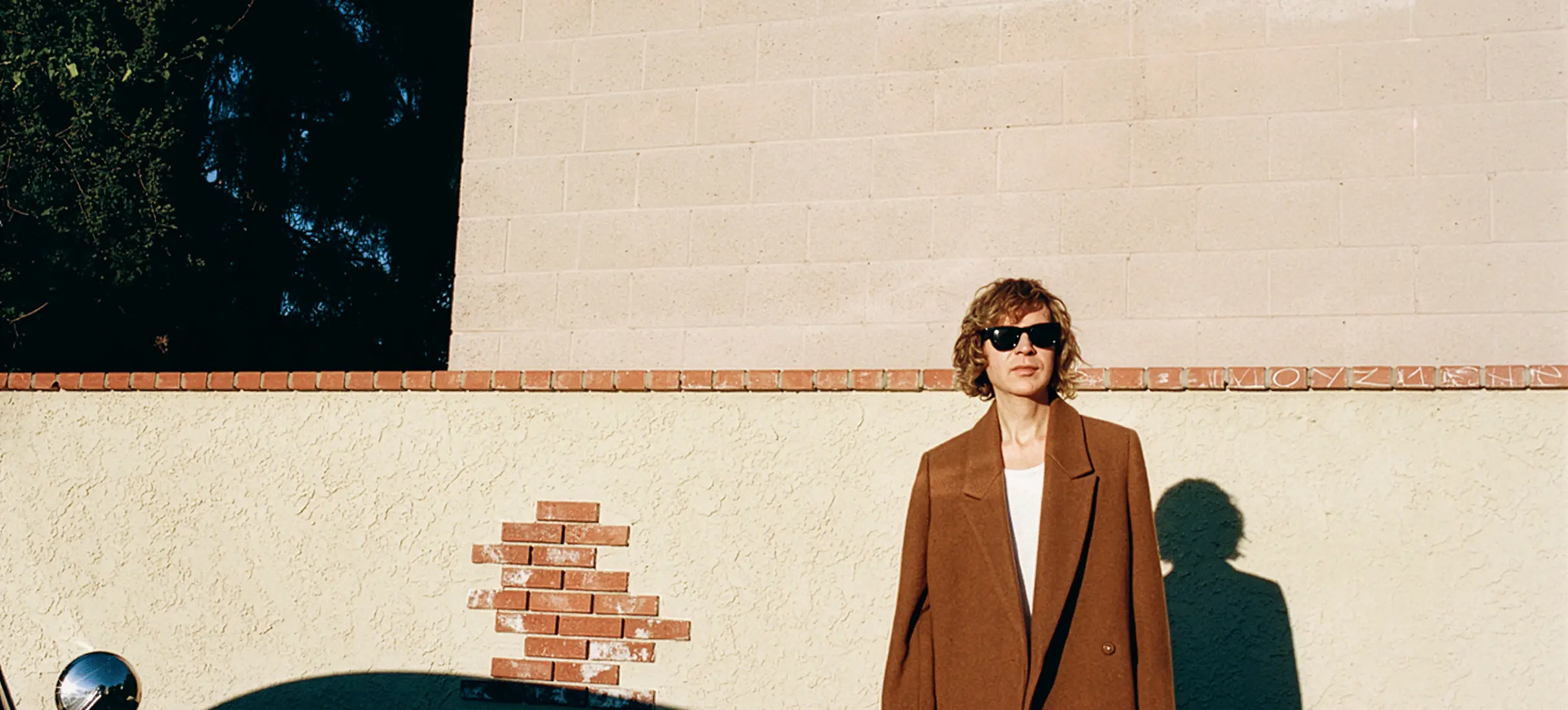 Person wearing brown coat and sunglasses standing against a beige wall with a shadow of a car in the foreground.