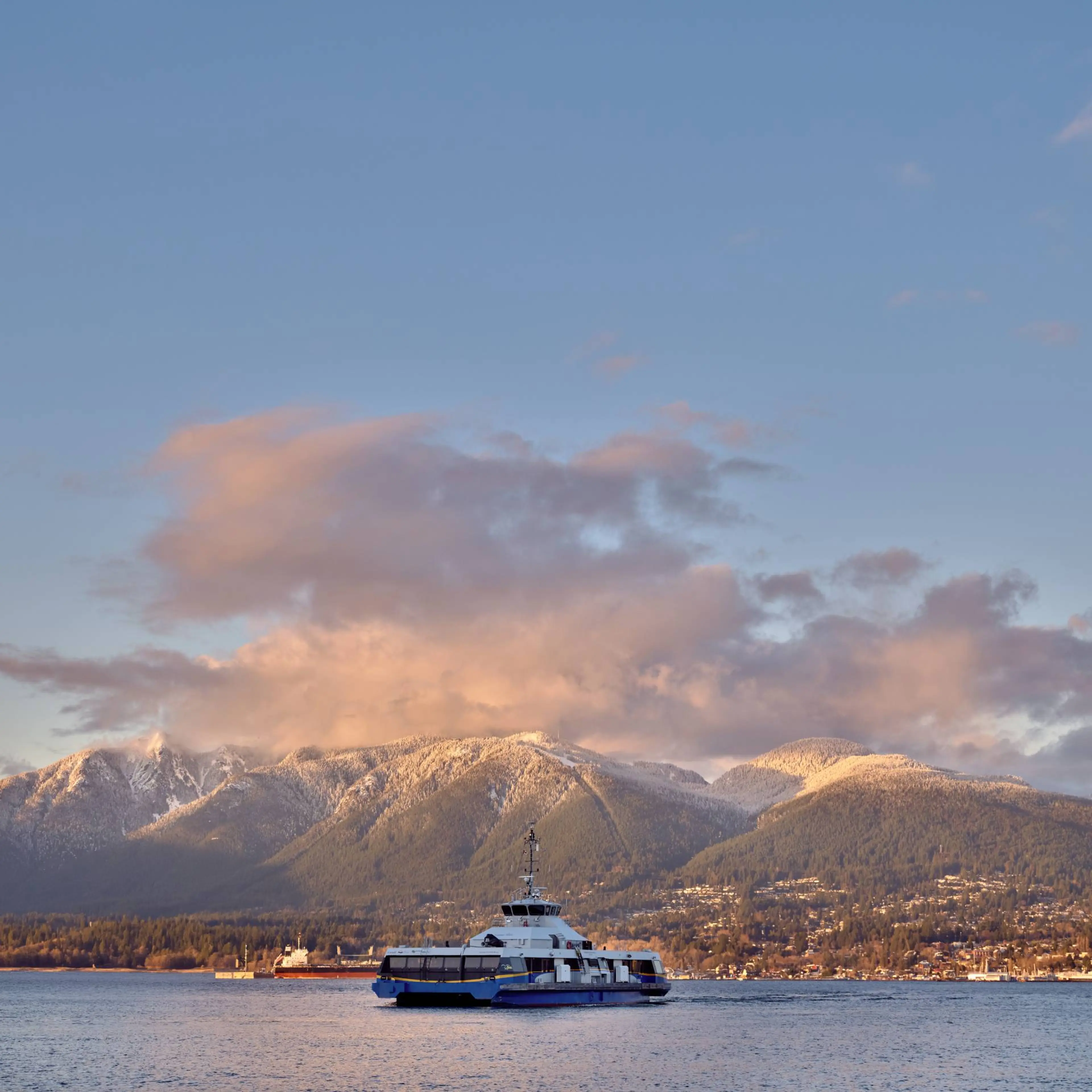 Ferry boat on calm water with snow-capped mountains and a cloudy sky at sunset in the background.