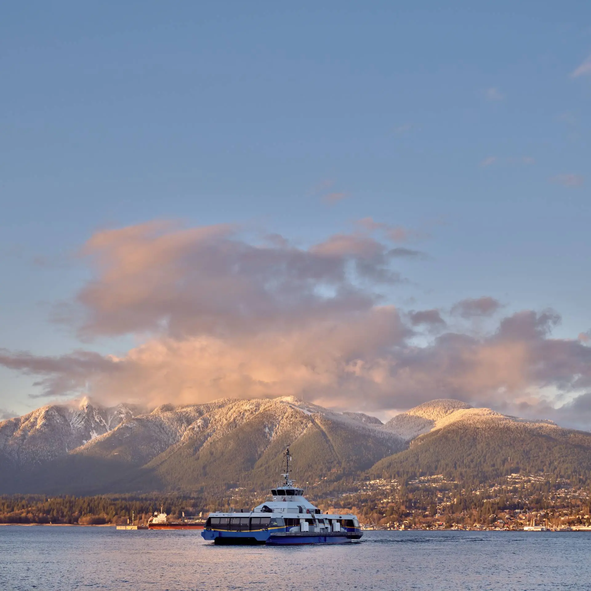 Ferry boat on calm water with snow-capped mountains and a cloudy sky at sunset in the background.