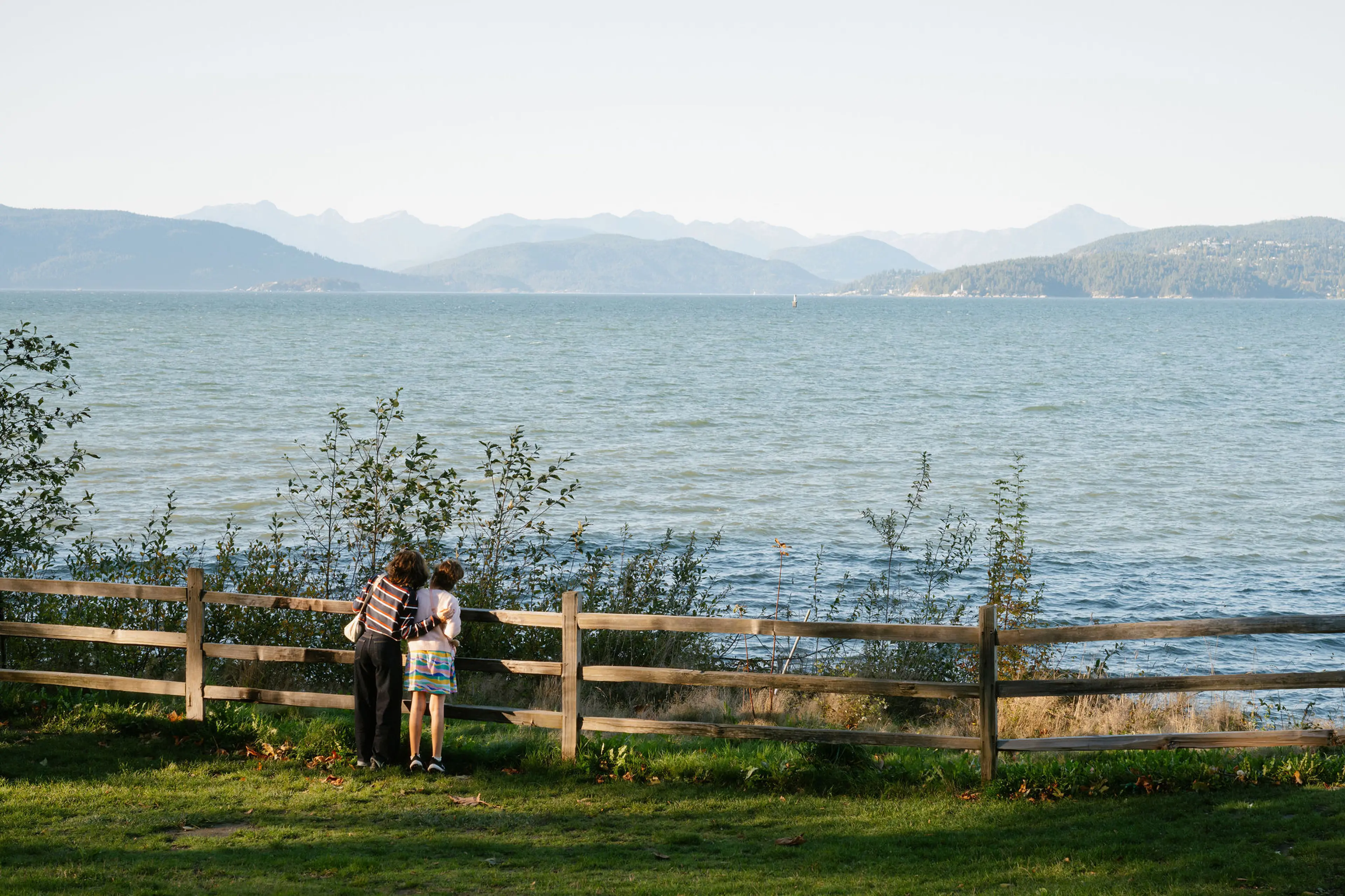 Two people standing by a wooden fence looking out over a large body of water with mountains in the distance.