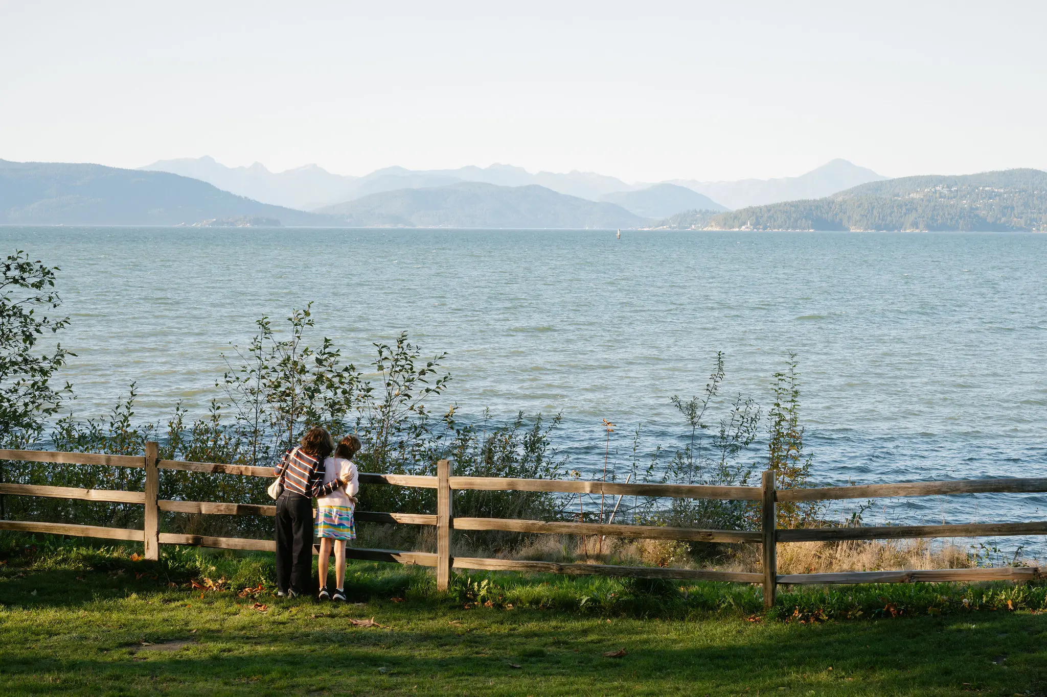 Two people standing by a wooden fence looking out over a large body of water with mountains in the distance.