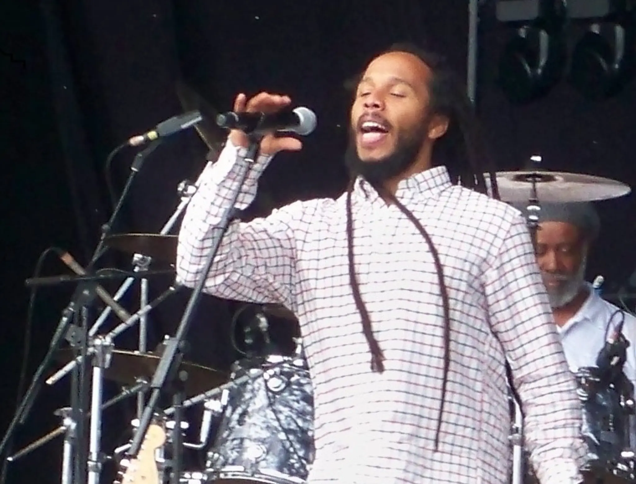 Man with long dreadlocks singing into a microphone on stage, wearing a checkered shirt and ripped jeans, with drums and guitars in the background.