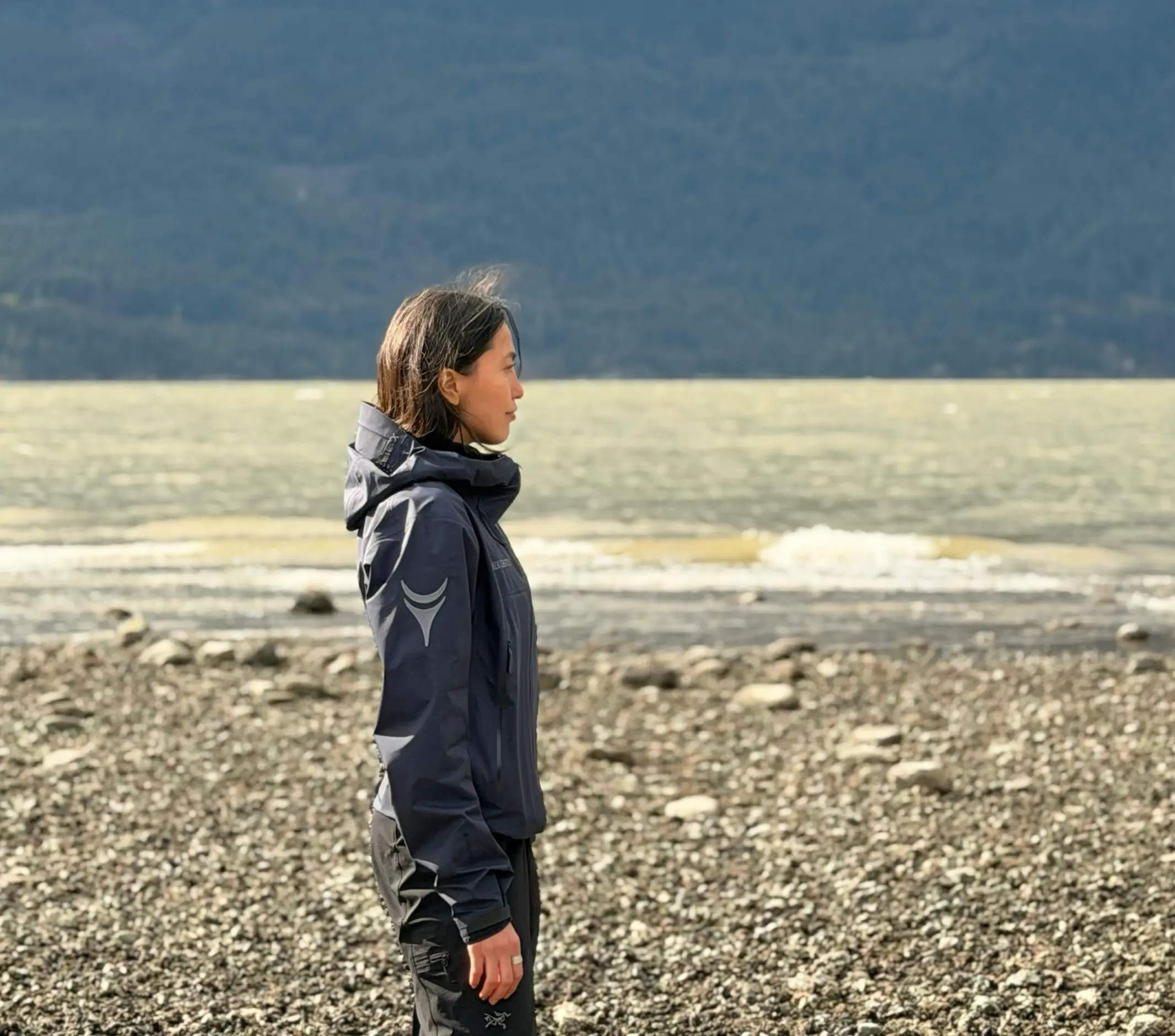 Person in a dark jacket standing on a rocky shore facing a body of water with mountains in the background.