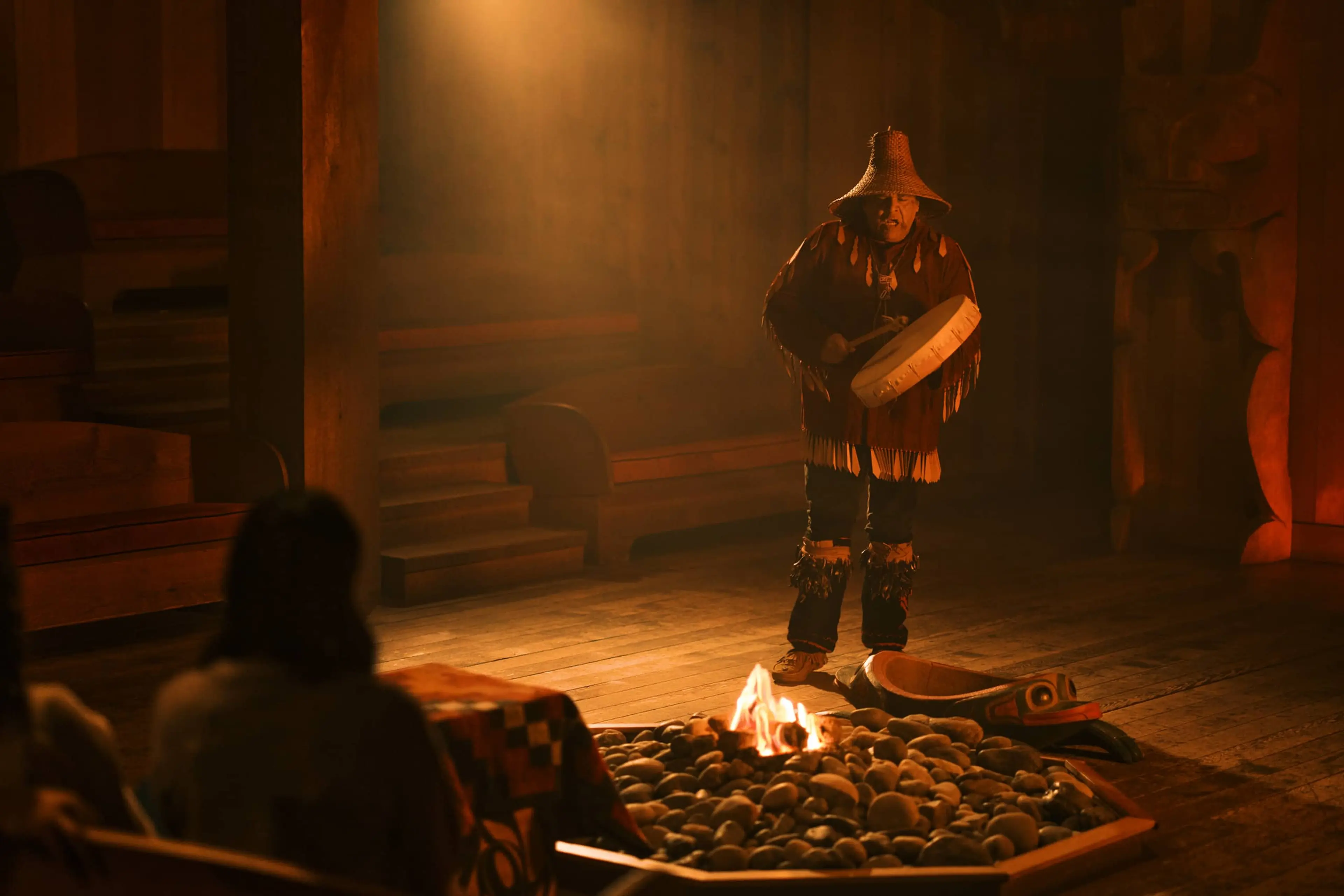 A person wearing traditional attire and a woven hat plays a drum near a fire pit inside a wooden structure with seated observers.