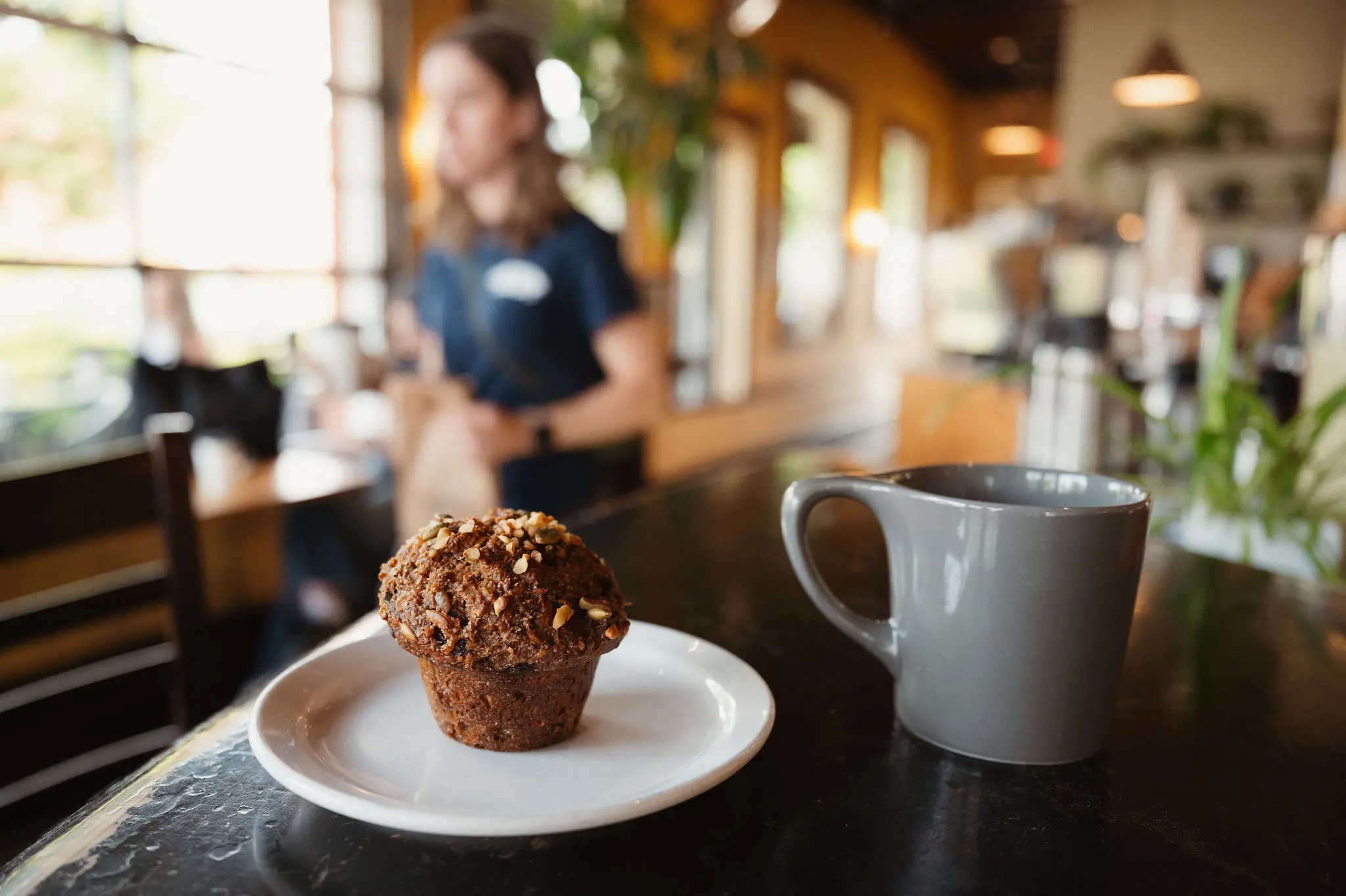 A muffin with nuts on a white plate next to a gray coffee mug on a dark table in a cafe with a blurred person in the background.