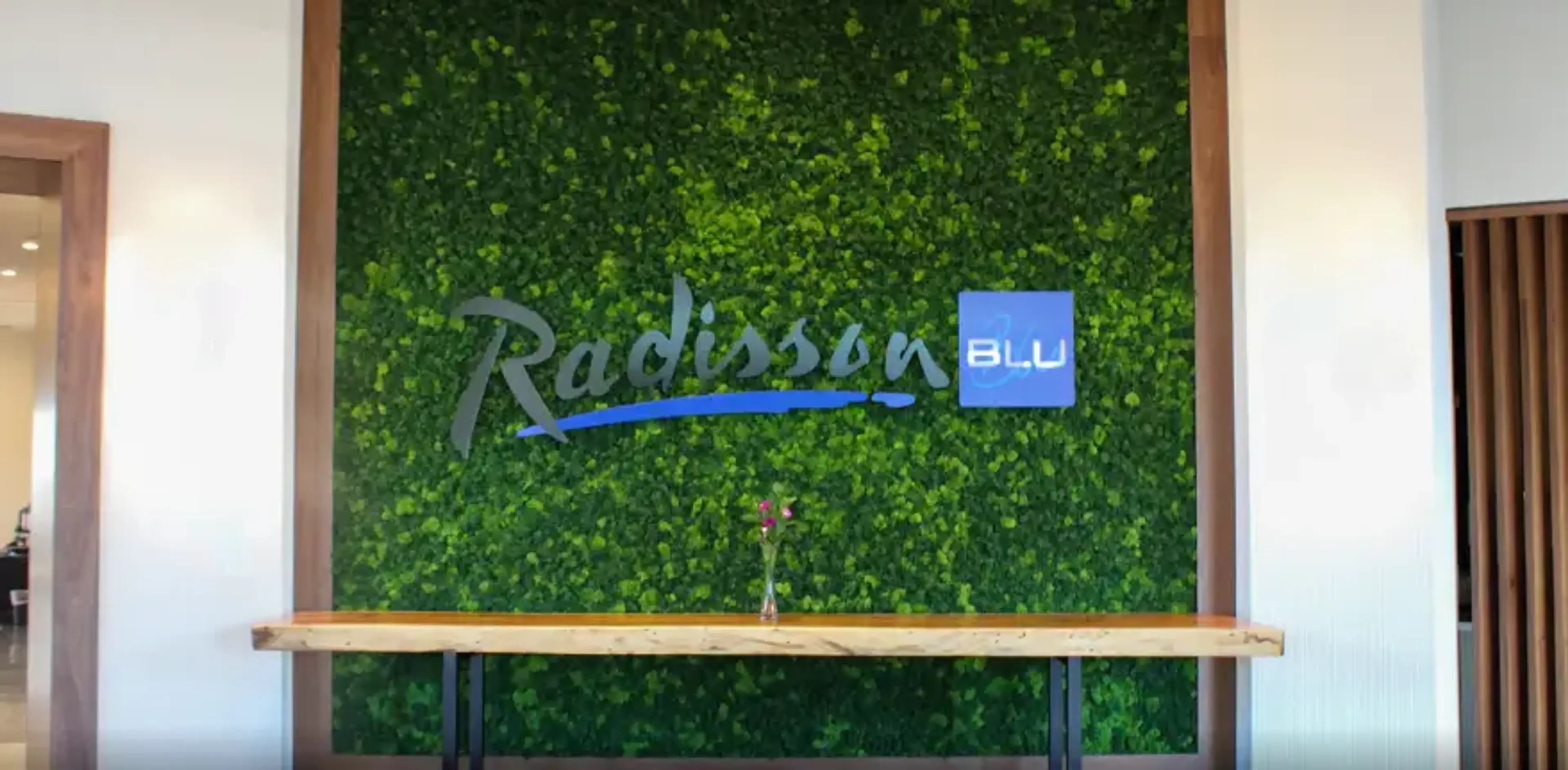 Green wall with Radisson Blu logo above a wooden table holding a small vase with flowers.