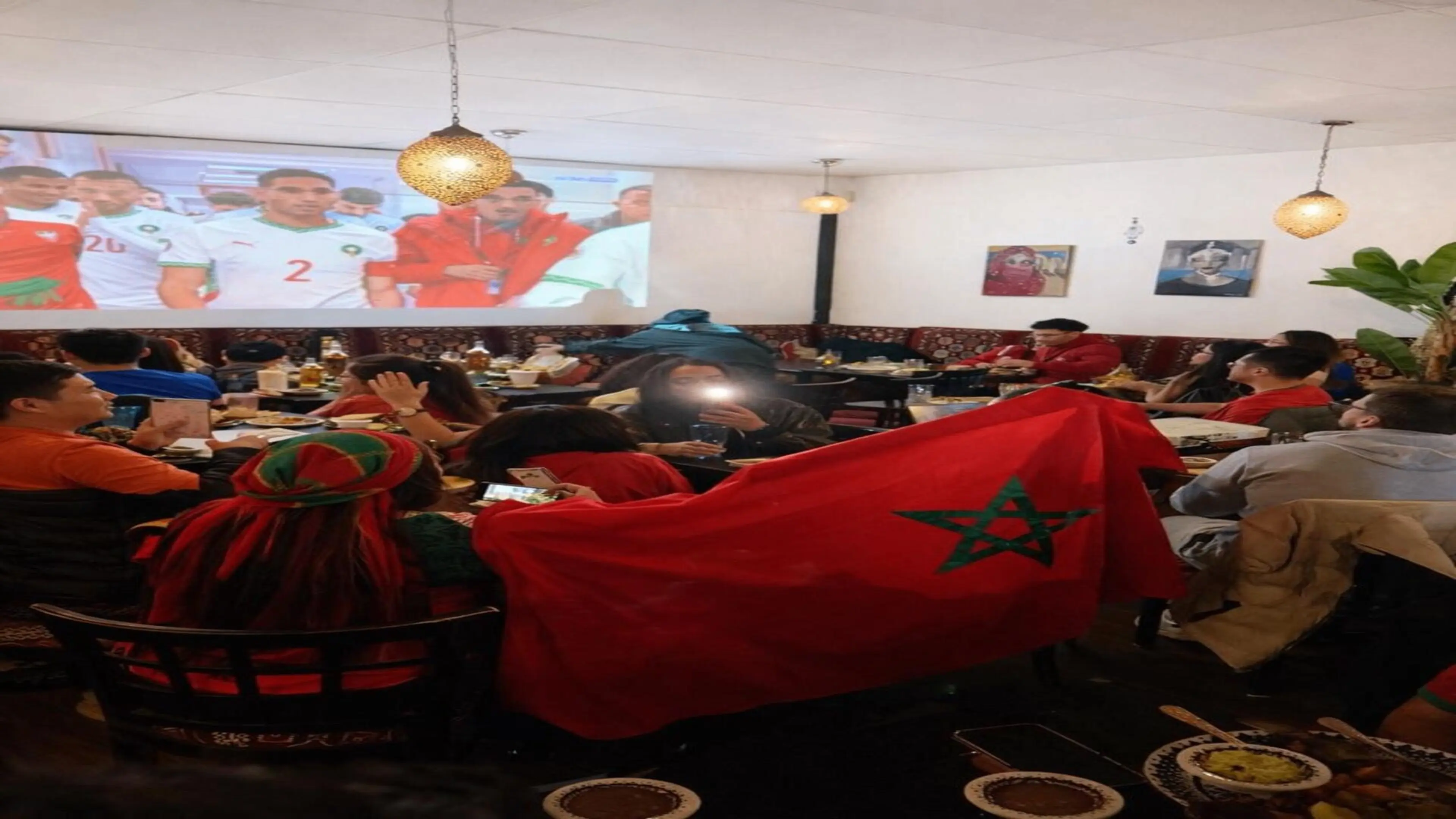 People in a restaurant watching a soccer match on a large screen, one person holding a Moroccan flag.