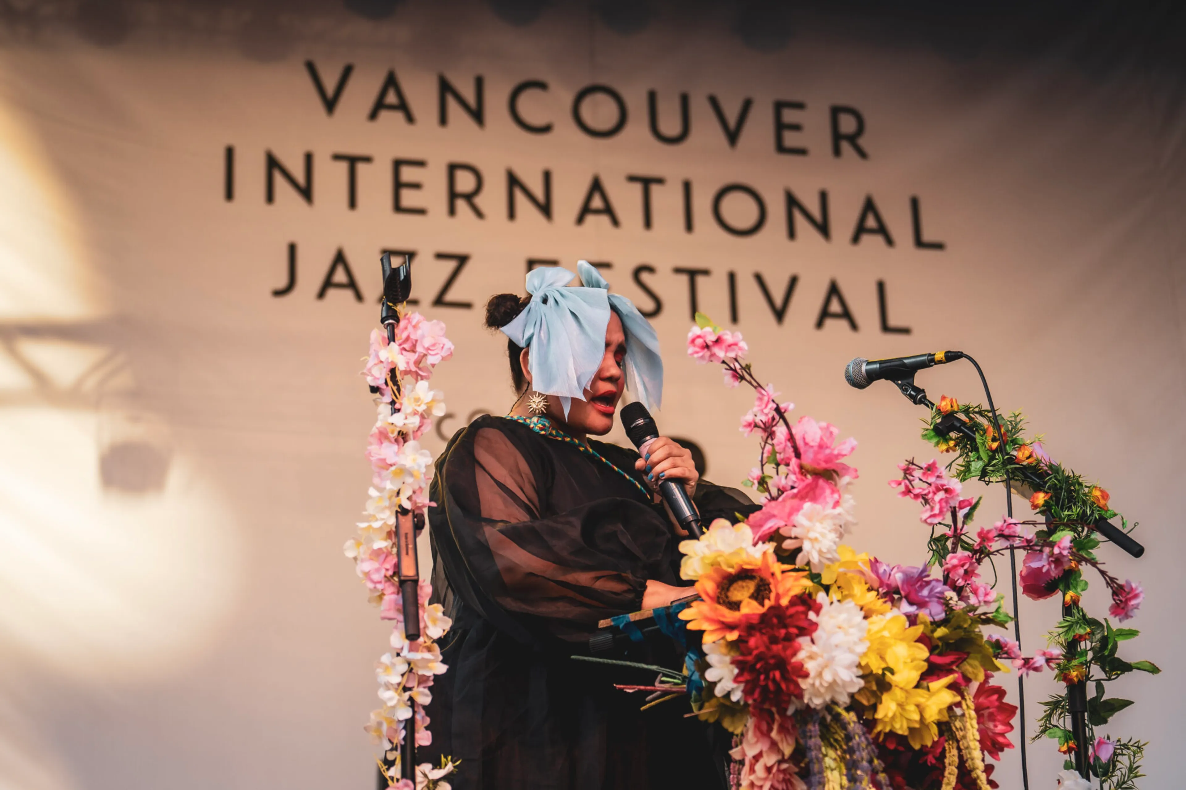 A person with a large blue bow on their head singing into a microphone on stage decorated with colorful flowers at the Vancouver International Jazz Festival.