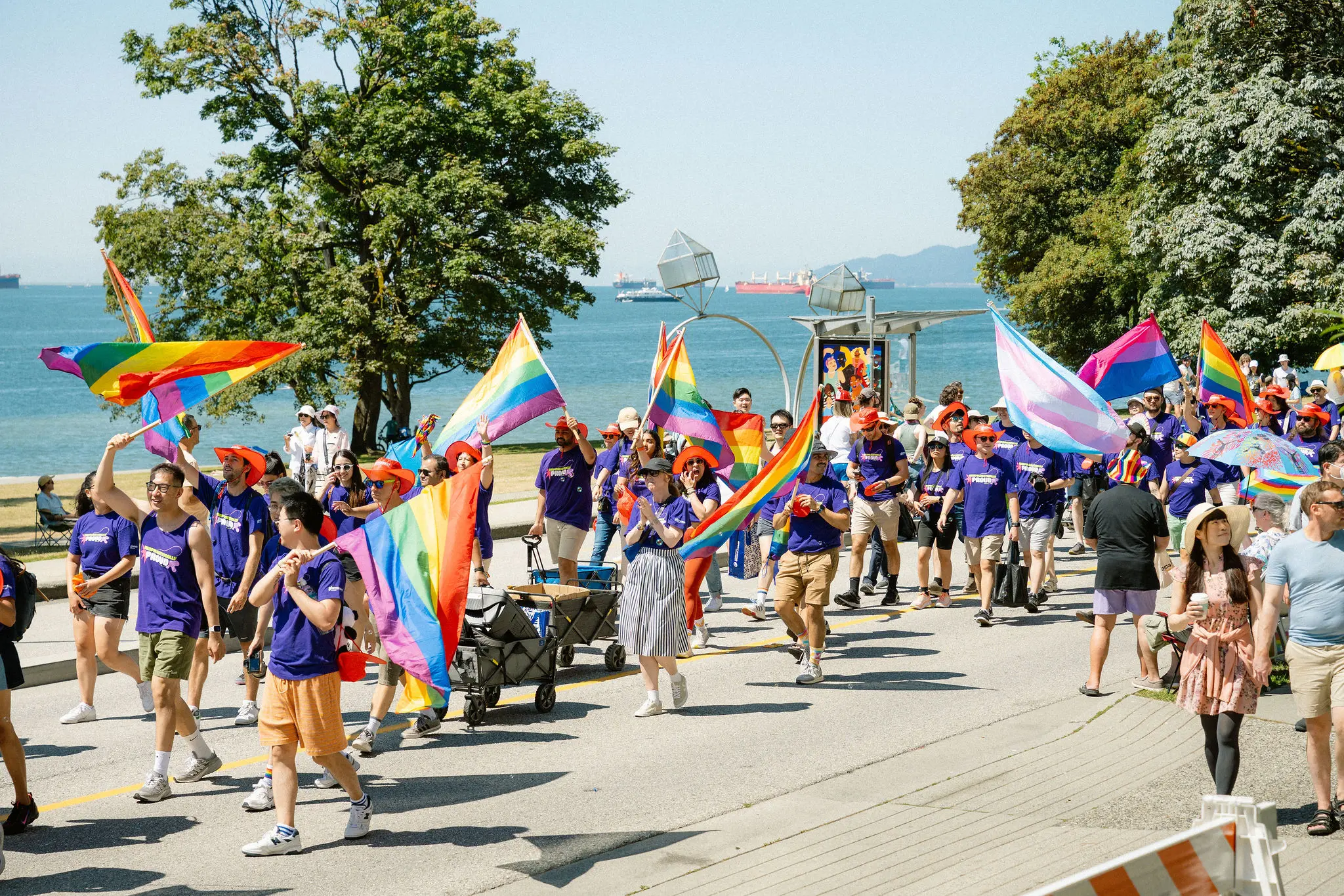Group of people walking outdoors near water, holding rainbow and transgender pride flags, wearing purple shirts and orange hats.