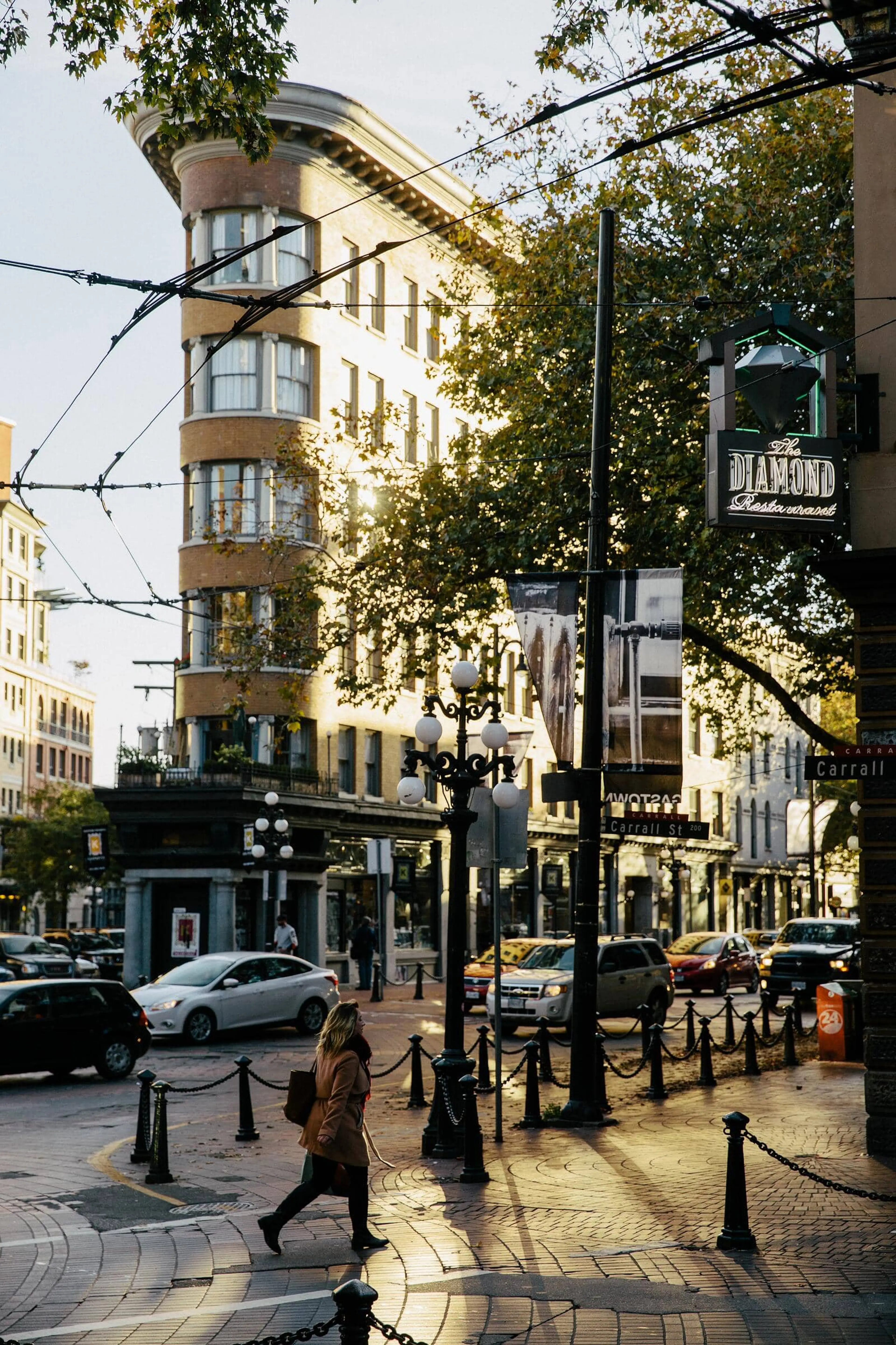 Person walking across a city street with a curved historic building and street lamps in the background during sunset.
