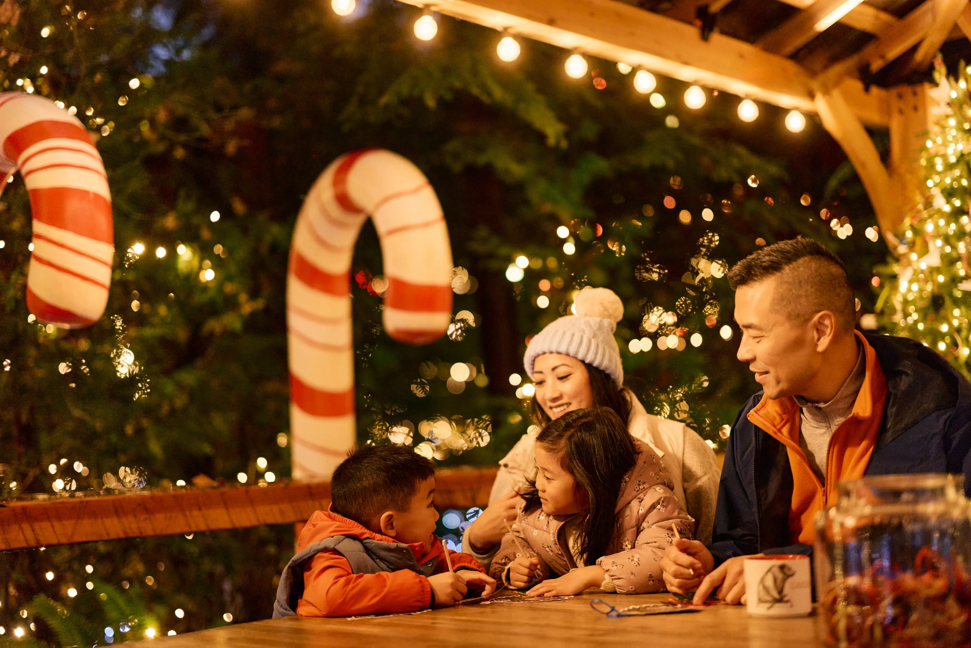 A family of four sitting at a wooden table outdoors at night, surrounded by festive lights and large candy cane decorations.