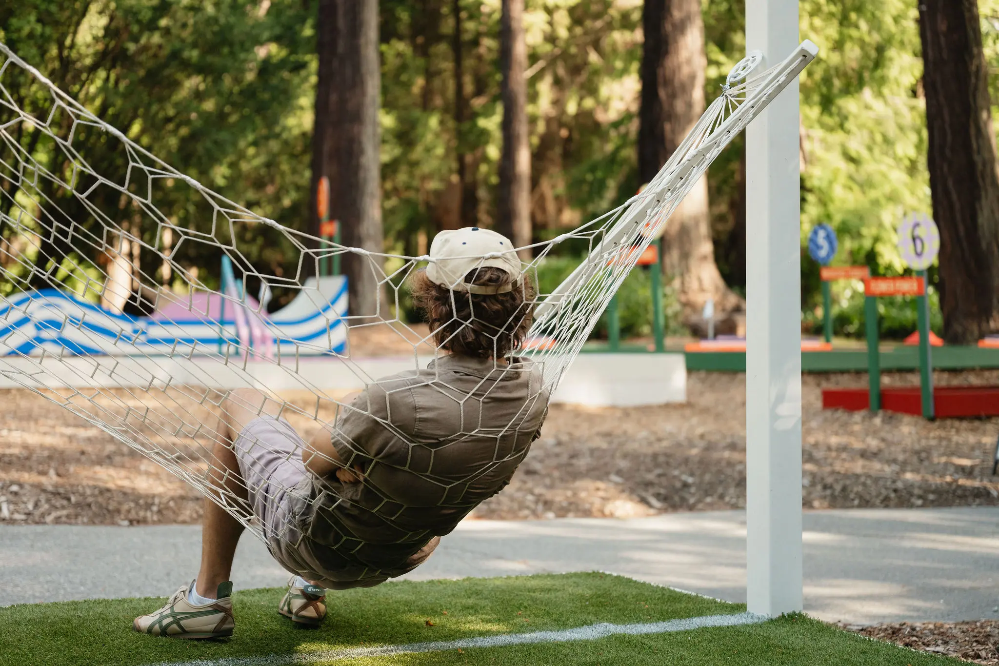 Person wearing a cap and casual clothes sitting in a net hammock outdoors with trees and mini golf field in the background.