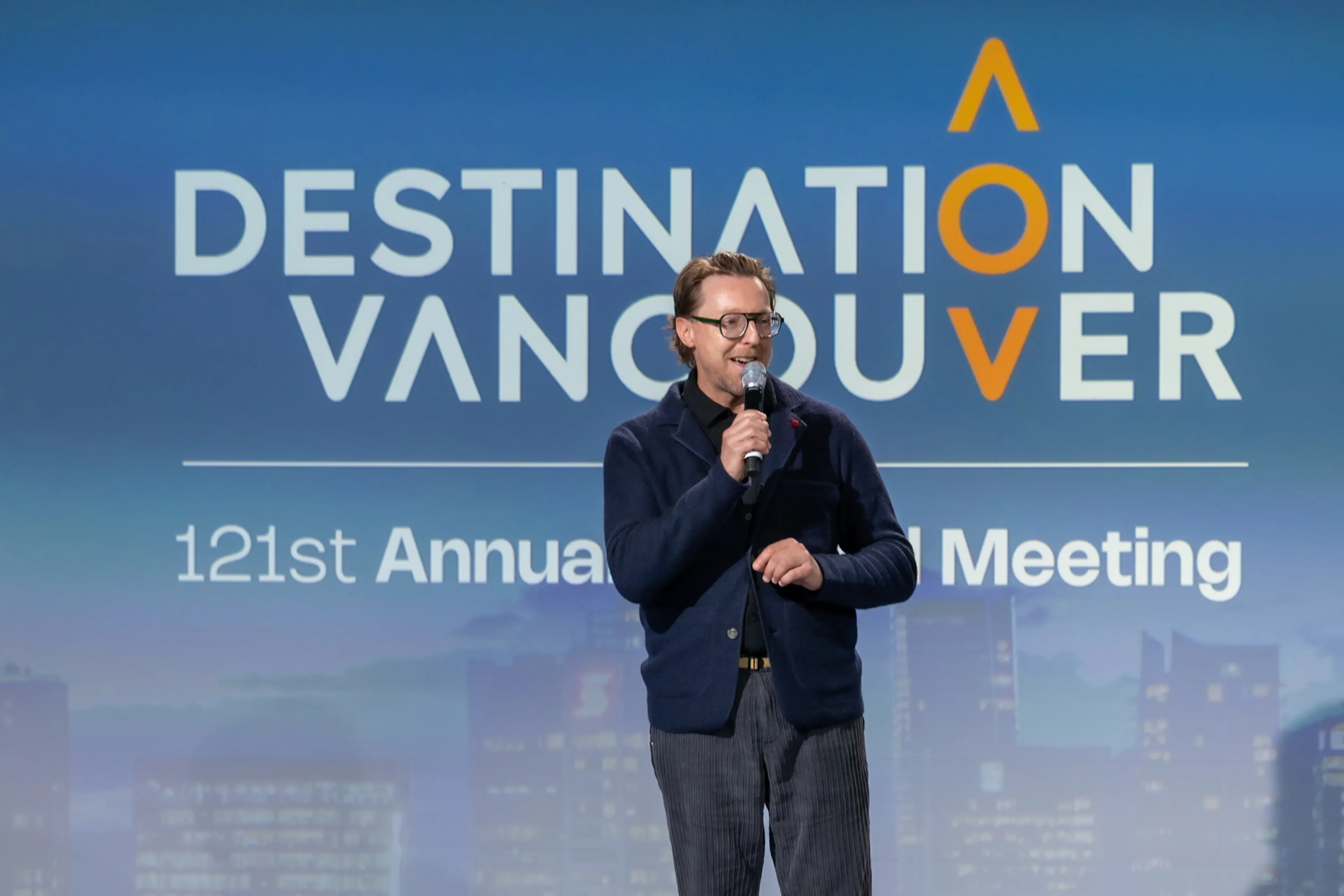 Person with glasses speaking into a microphone on stage with a backdrop reading 'Destination Vancouver 121st Annual Meeting'.